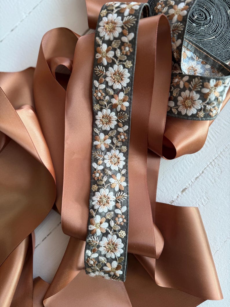 May include: A roll of brown satin ribbon with a floral embroidered trim. The trim features white flowers with gold accents on a dark green background.