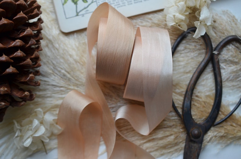 May include: A close-up of a spool of peach-colored satin ribbon. The ribbon is arranged in a loose coil and has a subtle sheen. The ribbon is lying on a soft, fuzzy surface.