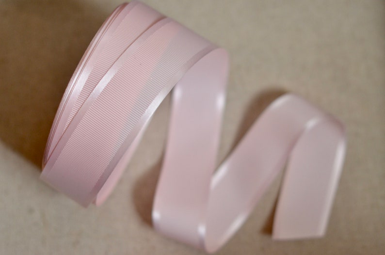 May include: A pale pink satin ribbon with a textured, ribbed surface. The ribbon is curled and folded, showing the sheen of the satin.