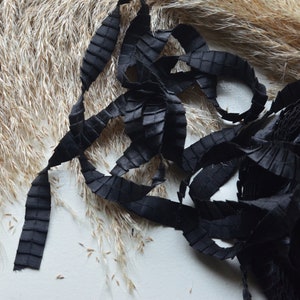 May include: Black pleated ribbon with a textured surface. The ribbon is arranged in a loose pile on a white background.