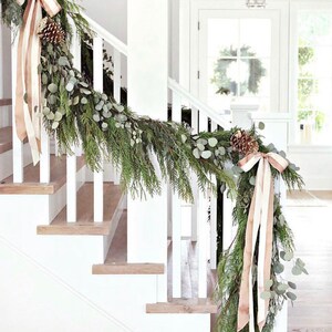 May include: A white staircase decorated with a green garland and beige ribbon. The garland is made of pine branches and eucalyptus leaves. There are pine cones and bows on the garland.