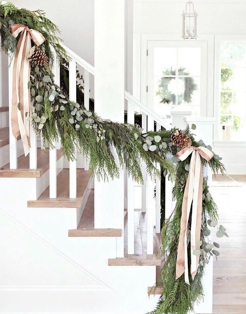 May include: A white staircase decorated with a green garland and beige ribbon. The garland is made of pine branches and eucalyptus leaves. There are pine cones and bows on the garland.