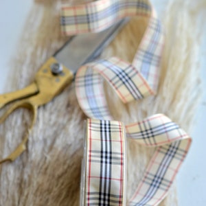 May include: A roll of beige ribbon with a black, white and red plaid pattern. The ribbon is lying on a fluffy white surface with a pair of gold scissors.