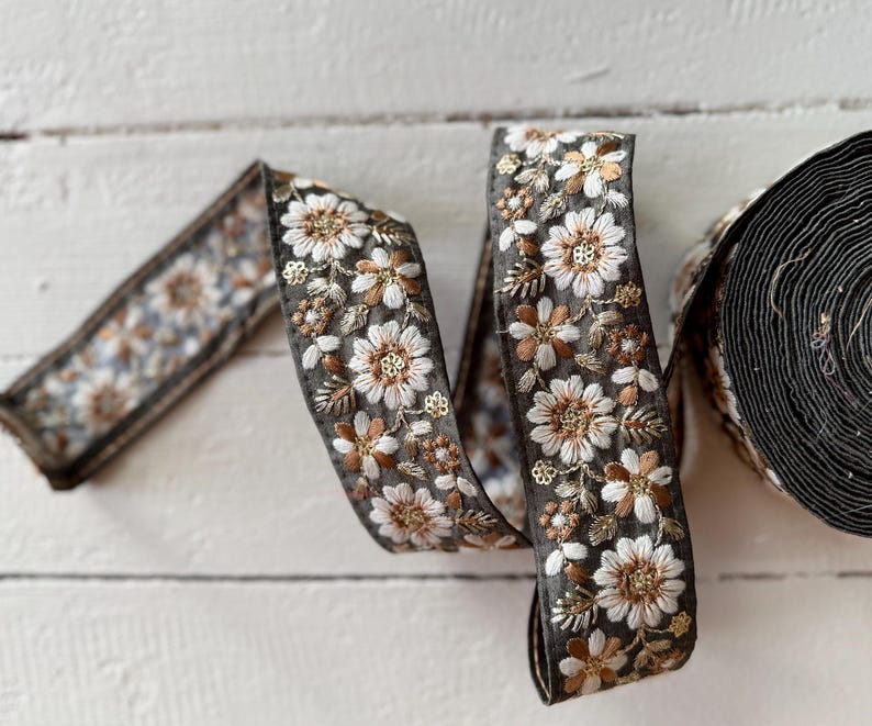 May include: A close-up of a black and white floral trim ribbon with gold accents. The ribbon is folded and twisted, showing the intricate details of the embroidery.