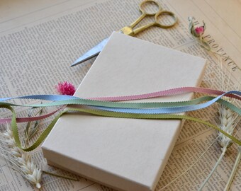 May include: A beige gift box with a light pink, light blue, and green ribbon tied around it. The box is sitting on a piece of paper with text.