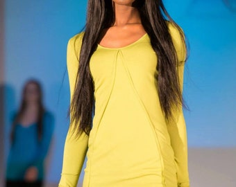 May include: A woman with long black hair is wearing a bright yellow long-sleeved shirt and black pants. She is standing on a runway with a blue background.
