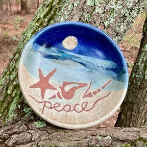 May include: A round ceramic dish featuring a coastal scene with a sandy beige foreground, a light blue shoreline, and a deep blue sky with a pale moon. A starfish and two seashells are depicted in brown, with the word peace written below them.