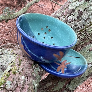 May include: A handmade ceramic colander and matching plate set. The colander is a deep blue with a turquoise interior and drainage holes. The plate is blue with a turquoise center and brown leaf designs. The set is resting on a tree branch.