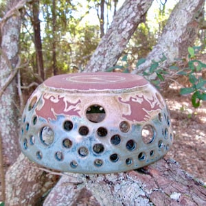 May include: A ceramic berry bowl featuring a rounded shape with numerous circular cutouts of varying sizes. The bowl has a two-tone glaze, with a reddish-brown upper section and a blue-toned lower section, and a circular stamp on the base.
