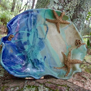 May include: A handcrafted ceramic serving dish with a coastal theme. The dish features a wavy, irregular shape and is painted with shades of blue, teal, and beige. Three starfish are attached to the surface, adding a tactile element.