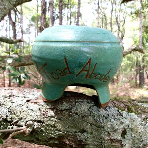 May include: A round, teal-colored ceramic toad abode with three short legs and a wide opening at the bottom. The words Toad Abode are etched into the front surface in a dark brown script. The ceramic has a smooth, glossy finish with subtle texture.