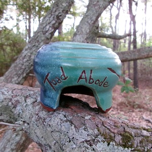May include: A handmade ceramic toad house with a turquoise and blue glaze. The words "Toad Abode" are carved into the side. The toad house is sitting on a tree branch.