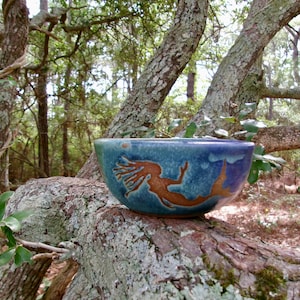 May include: A round ceramic bowl featuring a glazed finish in shades of blue and green. The exterior surface displays a prominent, embossed graphic of a mermaid in a warm brown tone, depicted with flowing hair and a curved tail extending across the side.