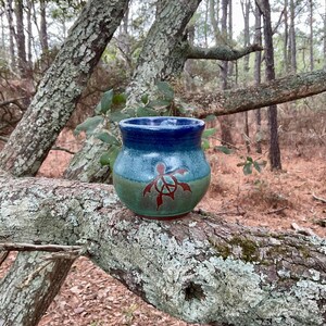 May include: A handmade ceramic mug with a blue rim, a turquoise body, and a green base. A red turtle design is painted on the front. The mug is sitting on a tree branch in a forest setting.