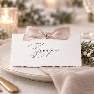 May include: A rectangular white place card with deckled edges featuring the name Georgie in black cursive script. A small, shimmering light brown ribbon is tied in a bow at the top centre of the card, which rests on a white ceramic plate.