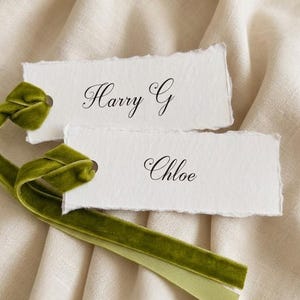 May include: Two rectangular white paper tags with deckled edges, each featuring black printed names. The top tag reads Harry G and the bottom tag reads Chloe. Each tag is attached to a long, soft olive green velvet ribbon loop.