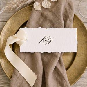 May include: A rectangular white place card with deckled edges featuring the name Katy in black cursive script. The card is tied with a cream-coloured satin ribbon and rests on a folded brown linen napkin placed atop a round brass-toned plate.