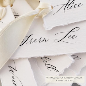 May include: A collection of white rectangular paper tags with deckled edges, each featuring black cursive calligraphy names. The tags are tied together with a cream-coloured silk ribbon. Text on the image reads: WITH MULTIPLE FONTS, RIBBON COLOURS & PAPER CHOICES.
