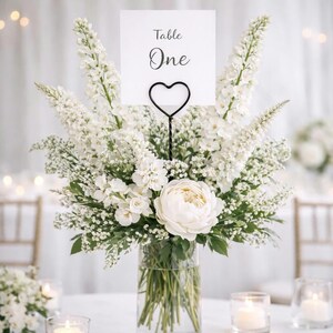 May include: A clear glass vase filled with a lush arrangement of white flowers including tall delphiniums, smaller filler flowers, and a large central peony. A black metal heart-shaped wire stand holds a white card printed with Table One.