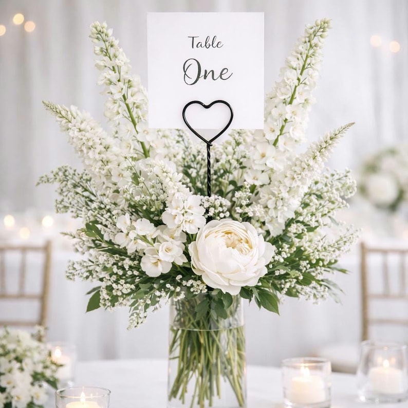 May include: A clear glass vase filled with a lush arrangement of white flowers including tall delphiniums, smaller filler flowers, and a large central peony. A black metal heart-shaped wire stand holds a white card printed with Table One.