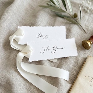 Puede incluir: Dos tarjetas de sitio rectangulares de papel blanco con bordes irregulares y caligrafía negra. La tarjeta superior dice Daisy y la inferior The Groom. Una cinta larga de satén blanco está enhebrada por el lado izquierdo de las tarjetas.