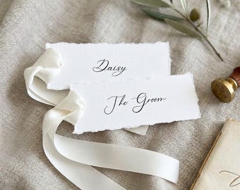 Könnte beinhalten: Zwei rechteckige weiße Papieranhänger mit Büttenrand und schwarzer Schreibschrift mit der Aufschrift Daisy und The Groom. Ein glattes weißes Satinband ist durch die linke Seite der Anhänger gezogen und bildet weiche Falten.