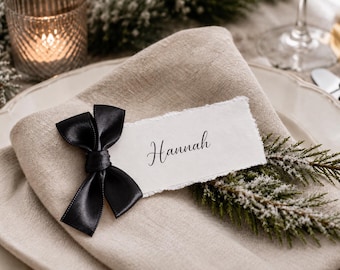 May include: A rectangular white place card with torn edges featuring the name Hannah in black script, adorned with a small black satin ribbon bow and a sprig of frosted evergreen, resting on a folded beige linen napkin.