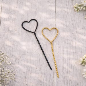 May include: Two heart-shaped wire sticks, one in matte black and one in metallic gold. Each stick features a twisted wire stem leading to an open heart outline at the top. The items are positioned diagonally, showcasing the thin, structured metal design.
