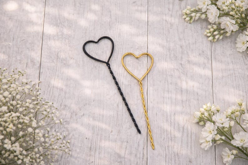 May include: Two heart-shaped wire sticks, one in matte black and one in metallic gold. Each stick features a twisted wire stem leading to an open heart outline at the top. The items are positioned diagonally, showcasing the thin, structured metal design.
