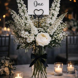 May include: A clear glass cylindrical vase filled with white flowers, including a large central peony and tall delphiniums. A black satin ribbon is tied around the vase. A white card with the text Table One is held by a black heart-shaped wire stand.