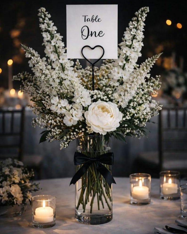 May include: A clear glass cylindrical vase filled with white flowers, including a large central peony and tall delphiniums. A black satin ribbon is tied around the vase. A white card with the text Table One is held by a black heart-shaped wire stand.