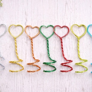May include: A collection of colorful wire heart-shaped place card holders. Each holder features a heart at the top, a twisted stem, and a spiral base. Colors include silver, gold, copper, green, red, yellow, and blue. Perfect for weddings or events.