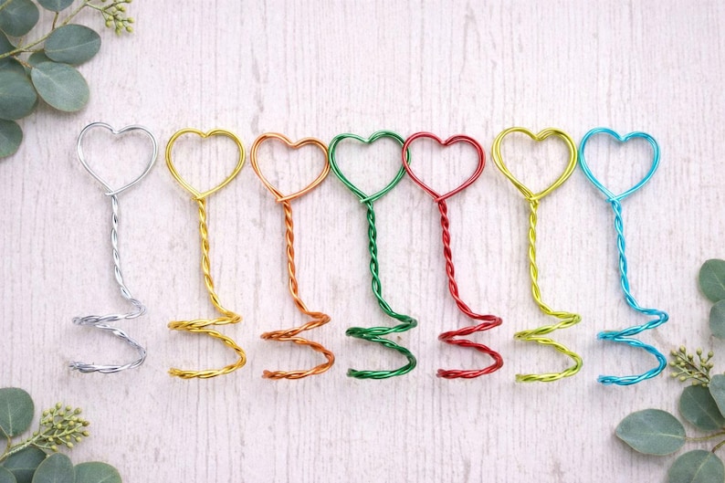 May include: A collection of colorful wire heart-shaped place card holders. Each holder features a heart at the top, a twisted stem, and a spiral base. Colors include silver, gold, copper, green, red, yellow, and blue. Perfect for weddings or events.