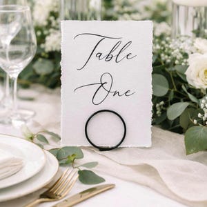 Op de afbeelding: Een witte tafelkaart met de woorden "Table One" in elegant zwart schrift. De kaart wordt vastgehouden door een zwarte metalen ring, geplaatst op een tafel met witte bloemen en borden met gouden randen. Thema: bruiloft of evenement.