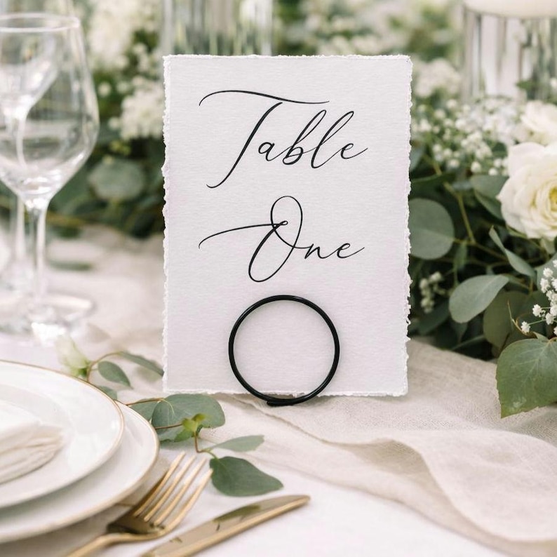 Op de afbeelding: Een witte tafelkaart met de woorden "Table One" in elegant zwart schrift. De kaart wordt vastgehouden door een zwarte metalen ring, geplaatst op een tafel met witte bloemen en borden met gouden randen. Thema: bruiloft of evenement.