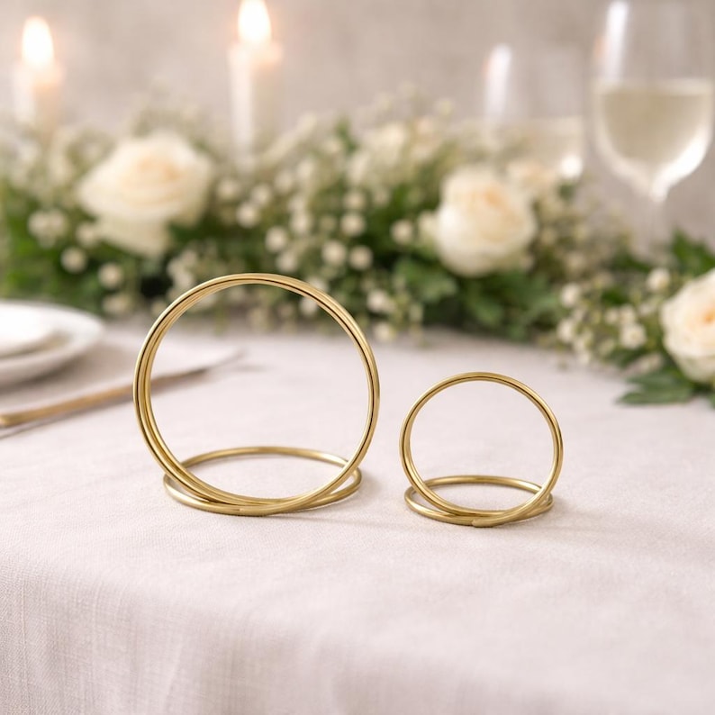 Op de afbeelding: Twee goudkleurige metalen ringvormige naambordhouders op een wit tafelkleed. De houders staan op de voorgrond, met een bloemstuk, kaarsen en glaswerk op de achtergrond. De ringen zijn verschillend van grootte.
