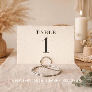 Puede incluir: Una tarjeta de número de mesa color crema con la palabra "TABLE" y el número "1" en negro. Un soporte de metal plateado sostiene la tarjeta. La imagen también incluye flores secas, velas y el texto "WEDDING TABLE NUMBER HOLDERS".