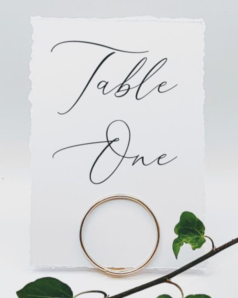Op de afbeelding: Een witte kaart met de woorden "Table One" in elegant zwart schrift. Een gouden ring houdt de kaart vast, met groene bladeren en een tak op de voorgrond. De kaart heeft een getextureerde, gescheurde rand.