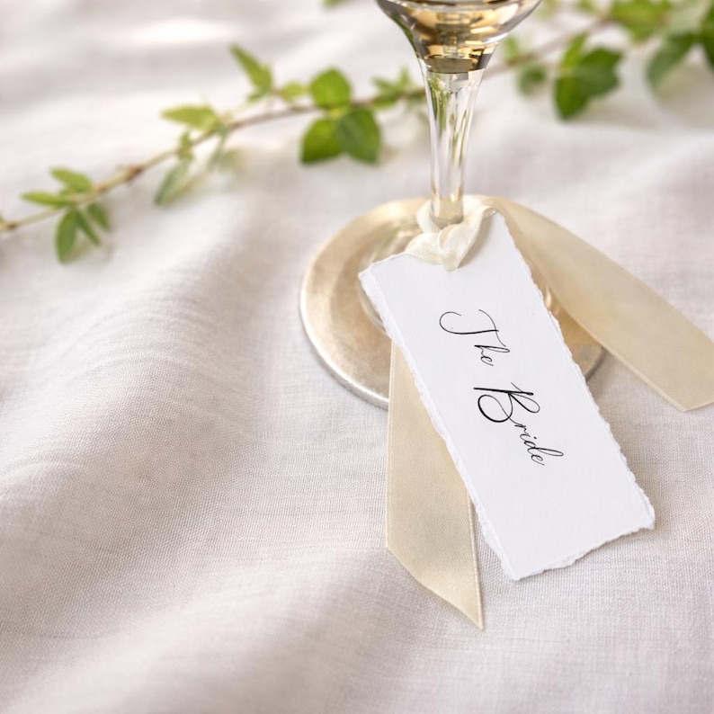 May include: A white rectangular paper tag with deckled edges featuring the black handwritten text The Bride. The tag is attached to the base of a clear glass stemware piece with a light cream-coloured satin ribbon tied in a simple knot.