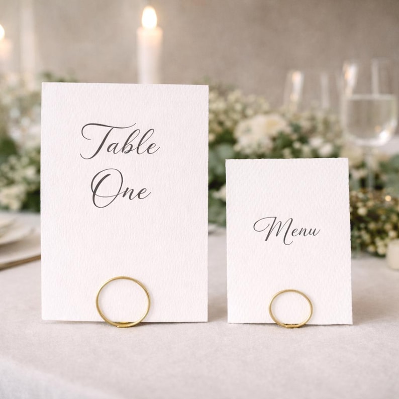 Op de afbeelding: Twee witte rechthoekige kaarten met elegante kalligrafie, &eacute;&eacute;n met de tekst "Table One" en de andere met "Menu". Elke kaart wordt vastgehouden door een kleine, goudkleurige ring. De kaarten liggen op een wit tafelkleed, met kaarsen en bloemstukken op de achtergrond.