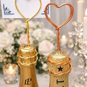 May include: Two champagne bottles with gold foil and heart-shaped wire toppers. One topper is gold, the other is copper. The bottles are on a table with candles and flowers in the background. The bottles have the labels "Laurent Perrier" and "Moet & Chandon".