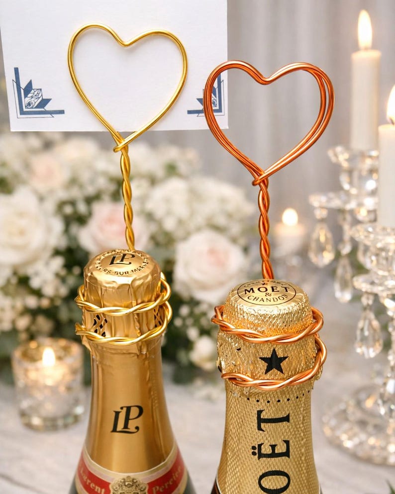 May include: Two champagne bottles with gold foil and heart-shaped wire toppers. One topper is gold, the other is copper. The bottles are on a table with candles and flowers in the background. The bottles have the labels "Laurent Perrier" and "Moet & Chandon".