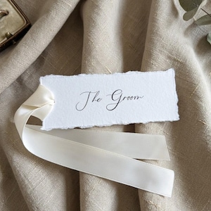 May include: A rectangular white paper tag with deckled edges featuring the black printed text The Groom in a cursive script font. The tag is attached to a smooth off-white satin ribbon that loops through a hole on the left side of the paper.
