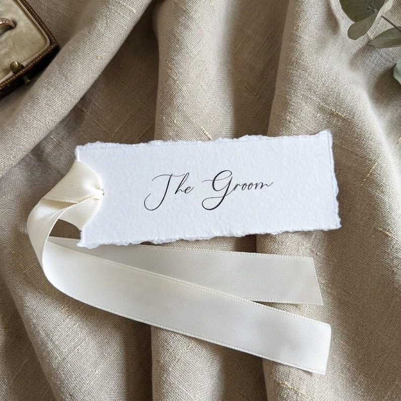 May include: A rectangular white paper tag with deckled edges featuring the black printed text The Groom in a cursive script font. The tag is attached to a smooth off-white satin ribbon that loops through a hole on the left side of the paper.