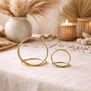 Op de afbeelding: Twee gouden metalen ringvormige kandelaars op een cr&egrave;mekleurig tafelkleed. De kandelaars zijn verschillend van grootte, met een grotere op de voorgrond en een kleinere erachter. De achtergrond omvat een vaas met gedroogde planten, een brandende kaars en andere decoratieve elementen.