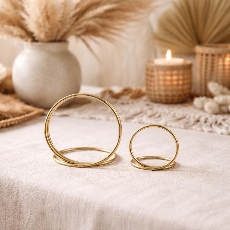 Op de afbeelding: Twee gouden metalen ringvormige kandelaars op een cr&egrave;mekleurig tafelkleed. De kandelaars zijn verschillend van grootte, met een grotere op de voorgrond en een kleinere erachter. De achtergrond omvat een vaas met gedroogde planten, een brandende kaars en andere decoratieve elementen.