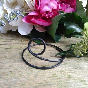 Puede incluir: Porta-bouquet de alambre metálico negro. El porta-bouquet está hecho de dos círculos entrelazados, con un círculo más pequeño en el centro.
