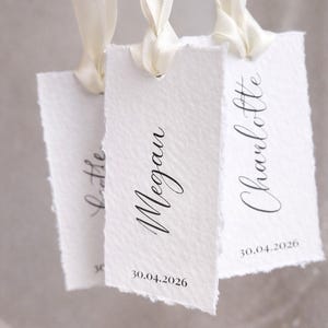 May include: Three rectangular white paper tags with deckled edges, each featuring a cream-coloured silk ribbon loop. The tags are printed with black script names including Megan and Charlotte, followed by the date 30.04.2026 in a simple font.
