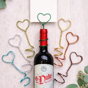 May include: A bottle of red wine with a red and green wrapped neck, surrounded by heart-shaped wire decorations in various colors. A white card is held by a green heart-shaped wire. The wine bottle label reads "The Duke Big Red Wine."