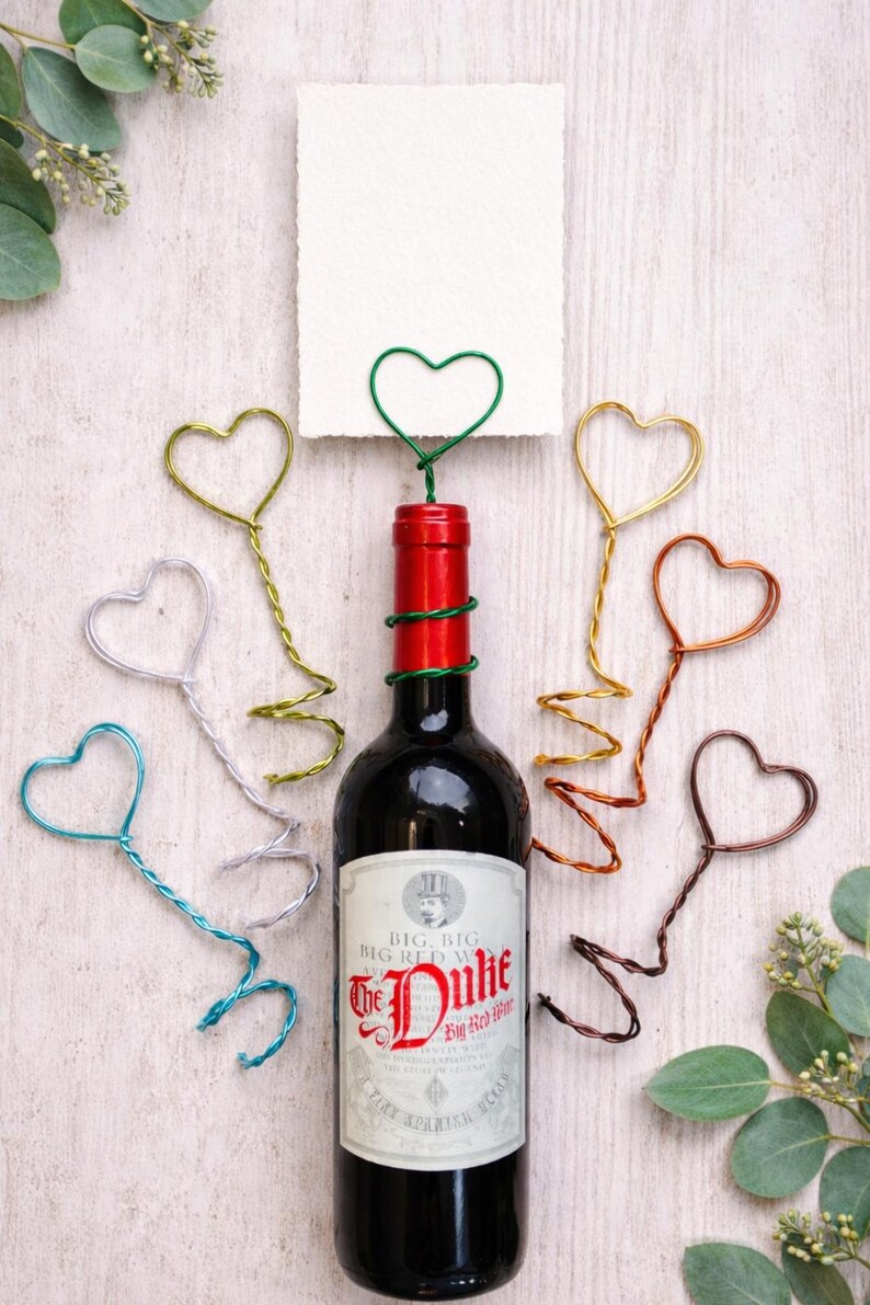 May include: A bottle of red wine with a red and green wrapped neck, surrounded by heart-shaped wire decorations in various colors. A white card is held by a green heart-shaped wire. The wine bottle label reads "The Duke Big Red Wine."
